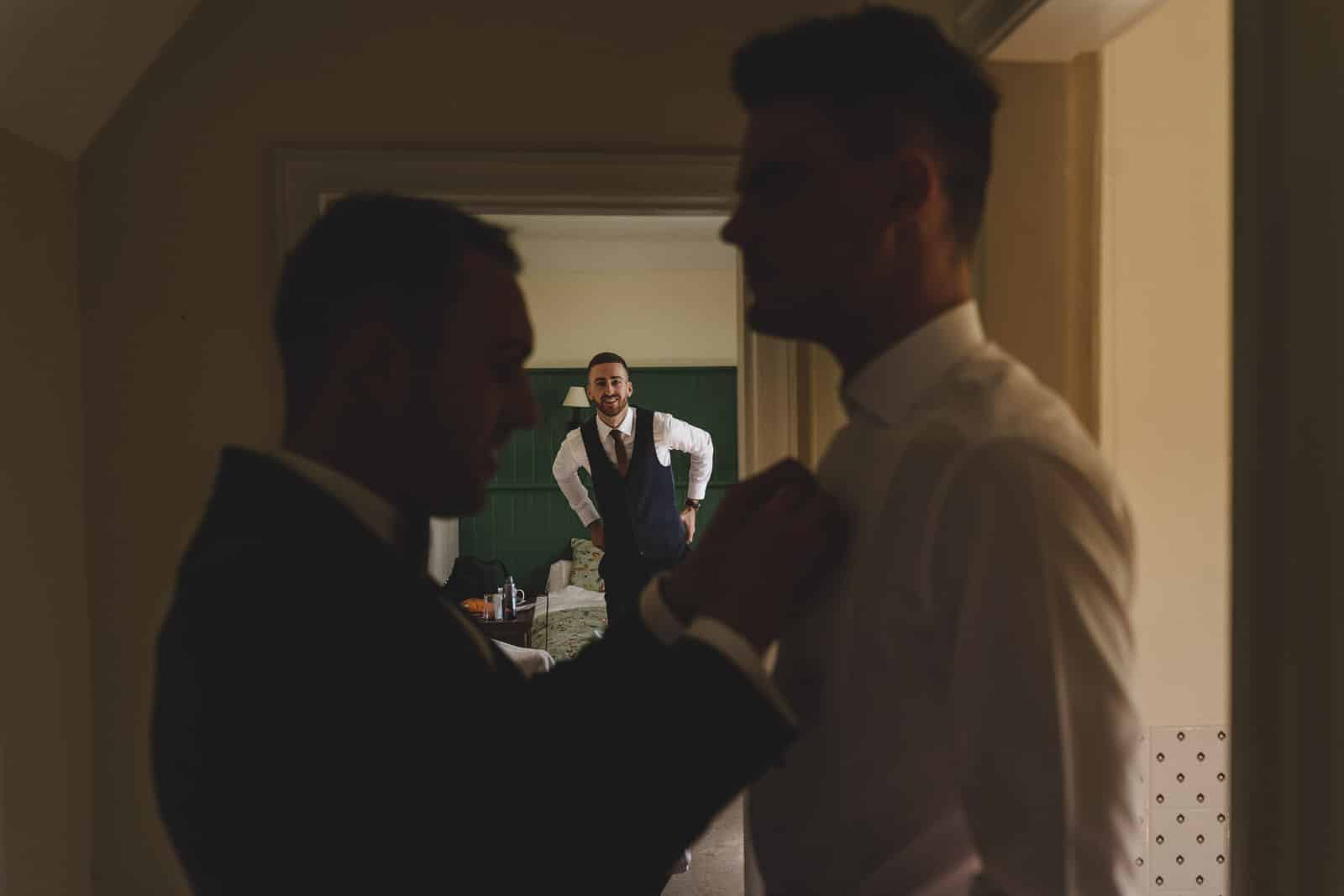 a groomsman appears through a door and smiles broadly at the groom and his best man
